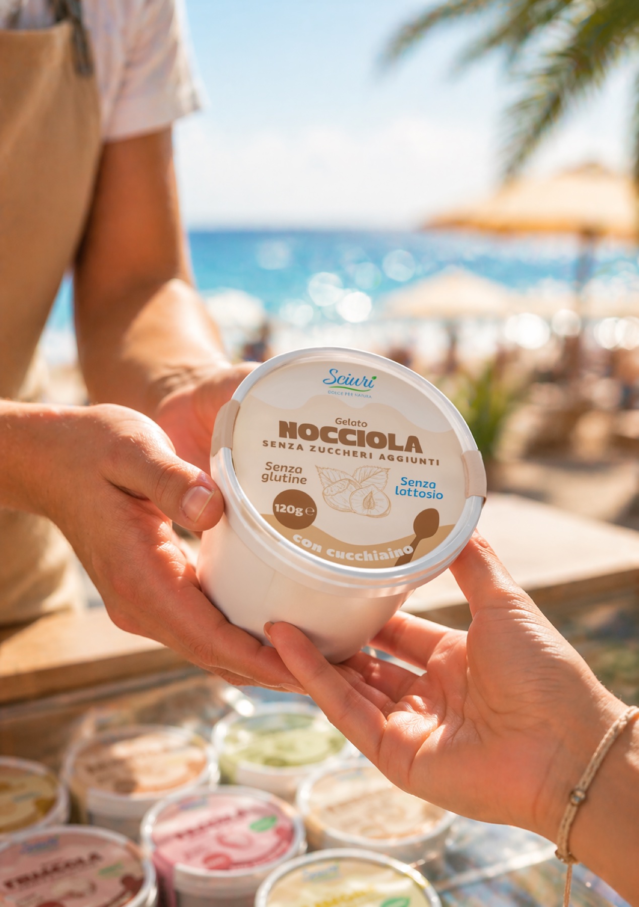 Due mani si passano una coppetta Sciurì alla nocciola su una spiaggia, con mare e ombrelloni sullo sfondo